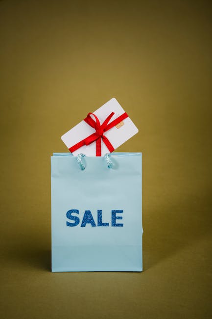 A light blue paper shopping bag with the word 'SALE' written in bold, dark blue letters is positioned upright on a plain, neutral background. Attached to the bag are two transparent plastic handles, and inside the bag is a white rectangular box tied with a red ribbon forming a bow on top. The box appears to be a gift or promotional item. The setting suggests a commercial or retail environment, but the focus is solely on the bag and gift box, with no other objects visible. This image could relate to house removals or packing and moving services provided by Man with Van Bow, especially in the context of planning a home relocation or packing process.