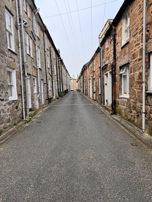 A narrow residential street in a suburban area with a gently descending road lined by white-painted houses and lush green trees on both sides. The pavement is partly shaded by overhanging branches, an