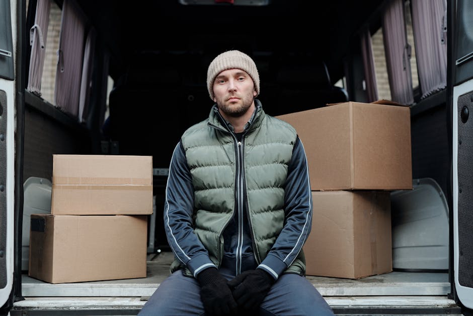 A man in a light grey knitted beanie, a puffer vest, and a dark blue jacket sits on the edge of an open van doorway during a home relocation process. The van's interior, visible behind him, contains several cardboard boxes of various sizes, some sealed with packing tape and others with visible flaps, arranged on the floor and near the sides. The boxes are made of standard brown corrugated cardboard and are stacked neatly, indicating they are packed for a moving service. The vehicle is parked on a paved surface, possibly a driveway or street, with a partial view of the house or building behind the van. The scene suggests a loading process involving furniture transport and packing materials, and the individual appears to be resting or waiting during the ongoing logistics of packing and moving, facilitated by [COMPANY_NAME], a professional removals service provider. The lighting is natural and even, highlighting the details of the packaging and the environment.