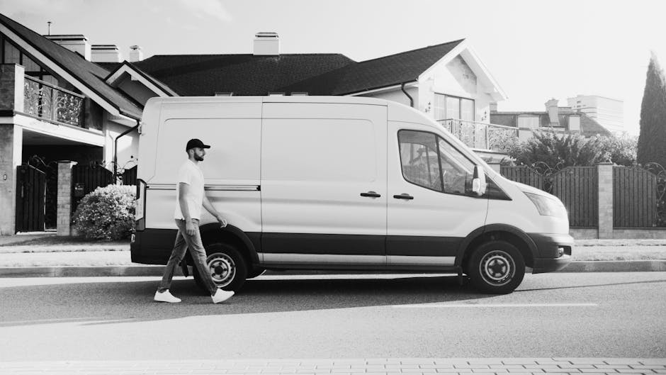 A man wearing a white t-shirt, jeans, and a cap is walking alongside a white panel van parked parallel to the pavement of a residential street. The van is positioned in front of a row of detached houses with pitched roofs, some featuring small balconies and fencing, characteristic of suburban properties. The vehicle's sliding door is closed, and it is equipped with standard side mirrors, hubcaps, and black bumpers. The man appears to be involved in a home relocation or furniture transport process, as the scene suggests a moving or packing activity typical of house removals, with the van ready for loading or unloading. The background includes trees, shrubs, and neighboring houses, and the overall lighting indicates daylight, with no visible signage or other objects in the vicinity. This image accurately depicts the moving logistics involved in professional removals, highlighting the vehicle, environment, and movement context relevant to [COMPANY_NAME].