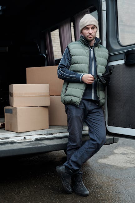 A man in a light grey knitted beanie, a puffer vest, and a dark blue jacket sits on the edge of an open van doorway during a home relocation process. The van's interior, visible behind him, contains several cardboard boxes of various sizes, some sealed with packing tape and others with visible flaps, arranged on the floor and near the sides. The boxes are made of standard brown corrugated cardboard and are stacked neatly, indicating they are packed for a moving service. The vehicle is parked on a paved surface, possibly a driveway or street, with a partial view of the house or building behind the van. The scene suggests a loading process involving furniture transport and packing materials, and the individual appears to be resting or waiting during the ongoing logistics of packing and moving, facilitated by [COMPANY_NAME], a professional removals service provider. The lighting is natural and even, highlighting the details of the packaging and the environment.